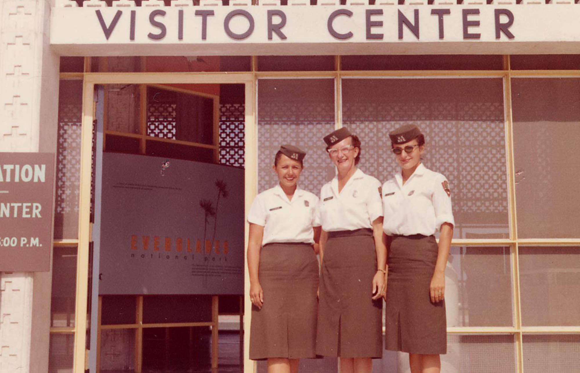 Three women stand in front of a visitor center building. They wear NPS uniform skirts, blouses, shield-shaped badges, and pillbox hats.